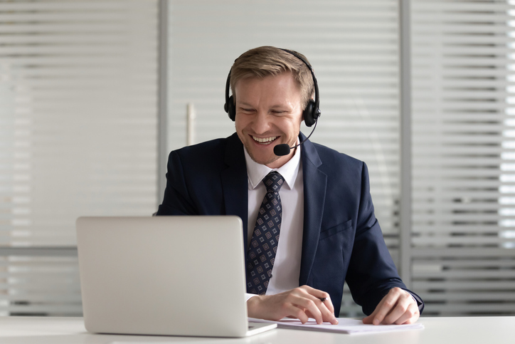 Smiling businessman in suit wearing headset laugh make notes having video call with business partners, happy male employee at workplace watch webinar or online course on laptop writing in notebook