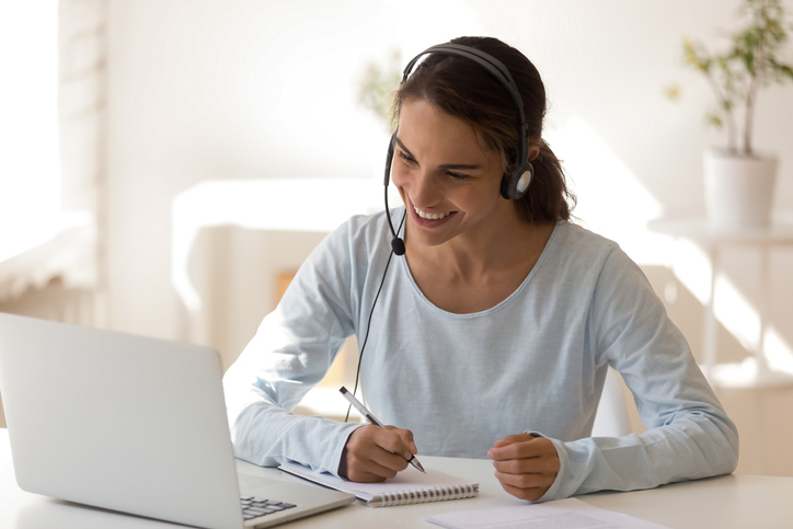 Happy mixed race young woman wearing headset, looking at laptop screen, watching educational lecture, enjoying interesting webinar, writing notes. Smiling employee holding video call with clients.