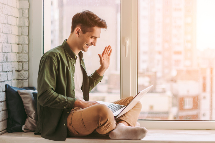 Happy stylish hipster man waving hand to laptop while having online video call at home. Young businessman freelancer sitting on windowsill while attend online meeting or conference. Social distancing