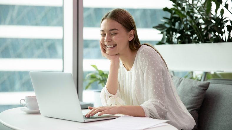 Smiling millennial Caucasian girl sit at desk laugh watching funny video or webinar on laptop, happy young female employee have fun browsing Internet on computer gadget, texting or messaging online