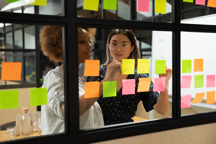 Concentrated african american young female scrum master working together with smart vietnamese businesswoman colleague, putting notes on sticky paper at window glass agile kanban board at office.