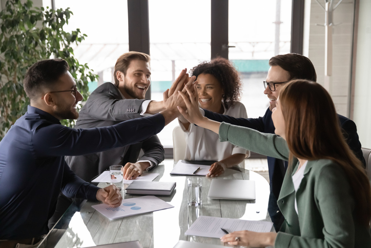 Overjoyed young mixed race business people sitting at table, joining hands in air, showing group unity, celebrating successful teamwork or professional achievement, raising team spirit at meeting.
