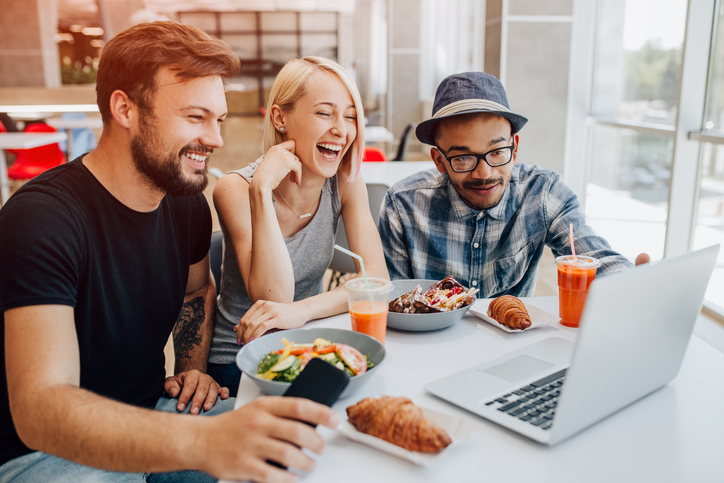 Excited diverse people laughing and using laptop to make video call while sitting at table and having lunch in cafeteria