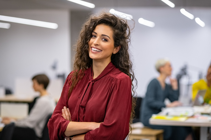 Successful businesswoman standing in creative office and looking at camera. Young latin woman entrepreneur in a coworking space smiling. Portrait of beautiful business woman standing in front of business team at modern agency with copy space.
