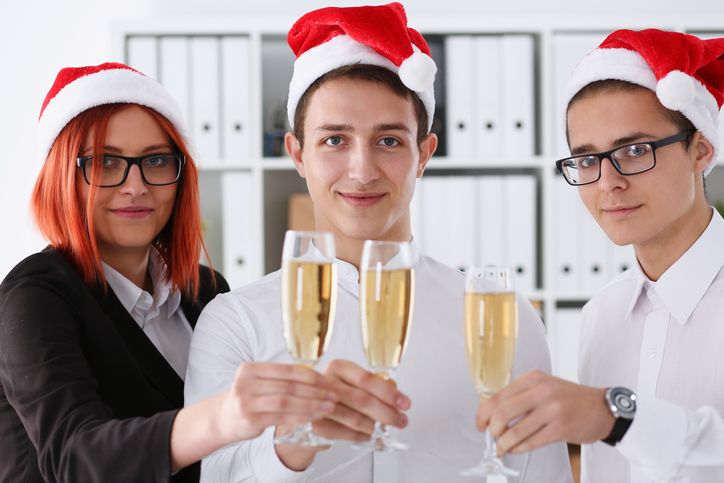 A group of businesspeople celebrating Christmas New Year corporate in the office while toasting holding glasses of champagne in hands on their heads wearing Santa cap.